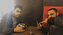 two men sitting at a table looking at their cell phones