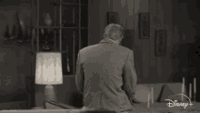 a man in a suit is sitting on a couch in a living room with a disney logo on the bottom right