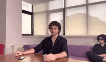a man in a black shirt is sitting at a table with a cup of coffee and a cell phone .
