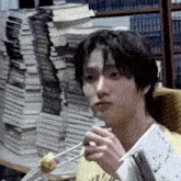a young man is sitting in front of a stack of books and eating food with chopsticks .