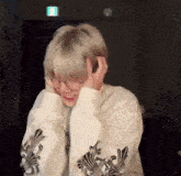 a person wearing glasses and a white sweater is covering their ears with their hands .