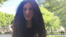 a woman with long curly hair is standing in front of trees