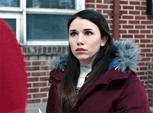 a woman wearing a burgundy jacket with a fur hood stands in front of a brick building