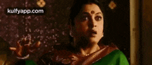 a woman in a green saree and a purple shawl is looking at the camera .