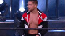 a shirtless wrestler in a red , white and black jacket is standing next to a railing .