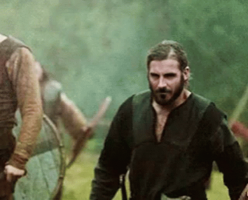 a man with a beard is walking in a field with a sword in his hand .