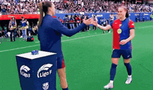 two women are shaking hands on a soccer field in front of a crowd .