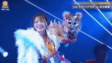 a woman in a feathered coat holds a mask in front of a blue background