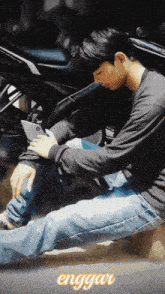 a man is sitting on the ground looking at his phone with the word enggau in the upper right corner