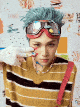 a young man with blue hair wearing goggles and a striped sweater is painting his face .
