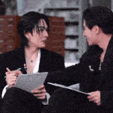 two men in suits are sitting next to each other and one is holding a clipboard and the other is holding a pen .
