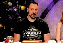 a man wearing an invulnerable vagrant t-shirt is sitting at a table