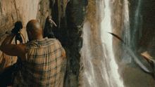a man in a plaid shirt holds a hammer while looking at a waterfall