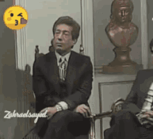 a man in a suit and tie is sitting in a chair with a statue in the background