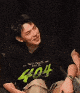 a man wearing a black shirt with the number 404 on it is sitting on a couch and smiling .