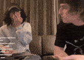 two men are sitting on a couch with one wearing a black shirt that says be nice