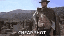 a man in a cowboy hat is standing in the desert holding a gun and a cape .
