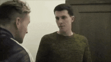 two men are standing next to each other and talking to each other . one of the men is wearing a green sweater .