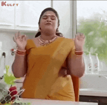 a woman in a yellow saree is standing in a kitchen with her hands up .