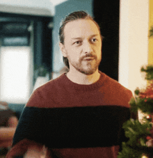 a man with a beard wearing a red and black striped sweater stands in front of a christmas tree