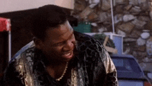 a man in a leopard print shirt and gold chain is sitting in front of a stone wall .