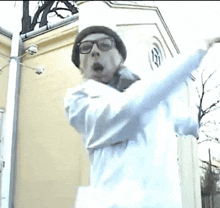 a man wearing glasses and a beanie is standing in front of a building with his arms outstretched