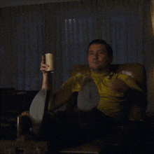 a man in a yellow shirt sits in a chair holding a cup and pointing