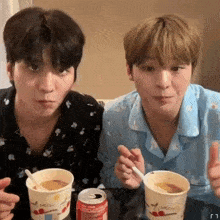 two young men are sitting at a table drinking from cups and a can of soda .