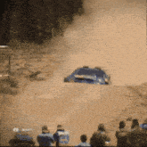 a blue subaru is driving down a dirt road with people watching