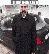 a man with a mustache is standing in front of a car with turk thinking written above him