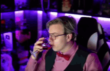 a man wearing glasses and a red bow tie is drinking a glass of wine while sitting in a chair .