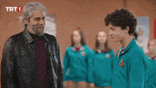 a man in a leather jacket stands next to a boy in a green shirt with trt1 on the bottom left