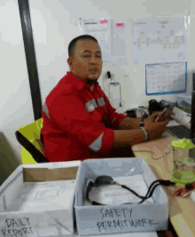 a man in a red shirt sits at a desk next to a box that says safety permit work