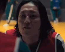 a man with long hair is wearing a red and white vest and looking at another man .