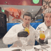 two men in white coats and black gloves are sitting at a table with drinks .