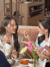 two women sit at a table with plates of food and flowers in a vase