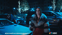a woman in a fur coat is standing in front of a white car with the nbc logo on the bottom