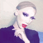 a drag queen with red lipstick and long eyelashes is taking a selfie with her hand on her chin .