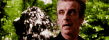 a man in a black shirt is standing in front of a tree and looking at the camera .