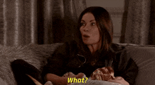 a woman sitting on a couch eating a sandwich and asking what