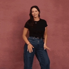 a woman in a black shirt and blue jeans is dancing in front of a pink wall