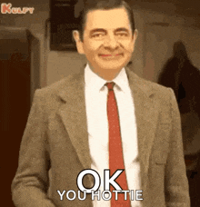 mr bean is wearing a suit and tie and smiling while saying `` ok you hottie '' .