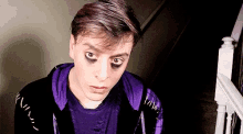 a young man wearing a purple shirt and a black jacket has black eye shadow on his eyes .
