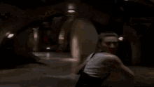 a woman in a white tank top is standing in a dark room and looking at something .