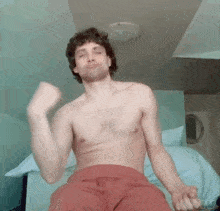 a shirtless man in red shorts is sitting on a bed with his arms in the air .
