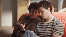 two men are sitting on a red couch looking at a cell phone