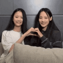 two young girls are sitting on a couch making a heart with their hands .