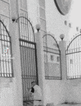 a black and white photo of a man standing in front of a building behind a fence .