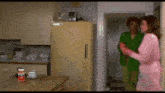 a woman in a pink robe stands in a kitchen next to a man in a green jacket