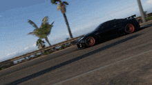 a black car with red wheels is driving down a road with palm trees in the background